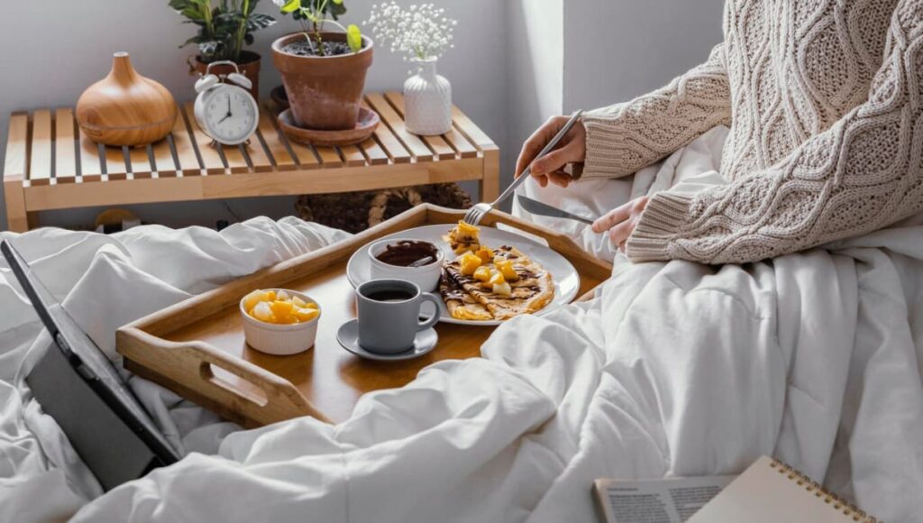 woman in a sweater cutting the pancakes with topping on the plate, a cup of coffee, and fruits