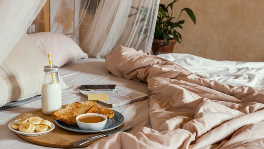 a plate with cut banana, bread, milk, and sauce on the bed with beige bedclothes