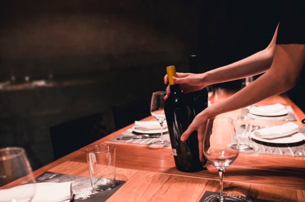 A person sets a table in restaurant, placing a dark bottle of wine next to clear wine glasses